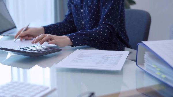 A female accountant with blue dotted blousy is using a laptop computer and calculator to calculate taxes at a glass desk in the office. Left side view.